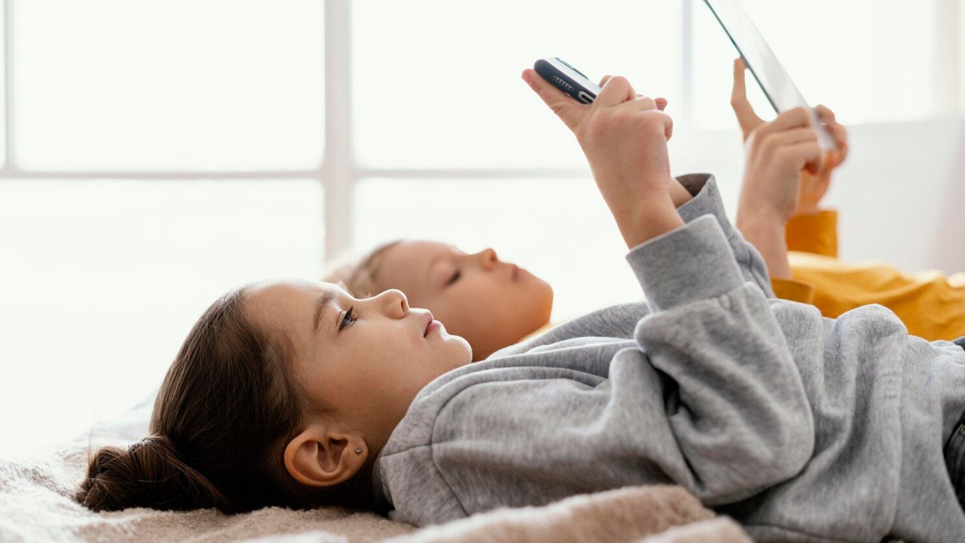 Niño y niña mirando el celular echados en la cama (Freepik)