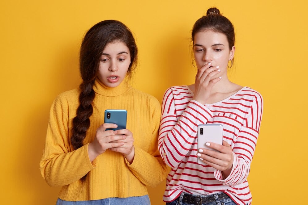 Dos mujeres jóvenes miran con sorpresa las pantallas de sus celulares (Freepik)